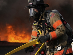 A firefighter in full protective equipment moves a yellow hose with fire in the background