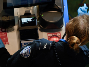 A TSA worker, seen over her shoulder, motions a passenger forward with a blue gloved finger
