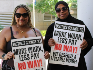 Two black women in sunglasses, one with a headscarf, hold signs saying ‘More work, less pay, NO WAY’