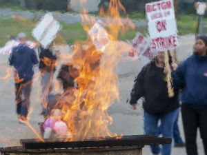 Workers holding picket signs that read BCTGM Local 3G On Strike march in the background, while a burn barrel with flames sits in the foreground.