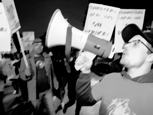 A man yells into a bullhorn with people in the background holding signs--is it blurry and black and white