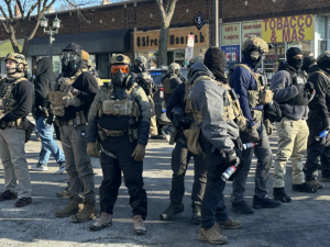 Agents covered head to toe in tactical gear and warm clothing stand in a diamond formation, facing outward, in a city street. Their faces are covered. The vests say "police" though they are not in uniform. They all wear heavy boots and some carry spray cans. Visible behind them is a one-story brick building lined with retail shops, with faded signs reading "8 Street Nourish" and "Tobacco & Mas."