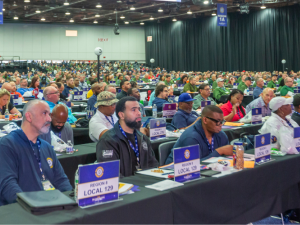 UAW delegates (men and women, Black and white) sit listening at the union's 2026 convention. Table signs identify those in the foreground as from Region 8, Locals 129 and 276.