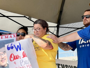 A woman cries while two men in blue Machinists t-shirts comfort her with a hand on her shoulder. Next to her is a woman holding a sign that says “Free Max.”