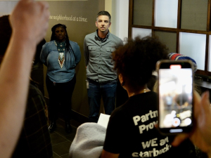 Two managers stand in a hallway as Starbucks workers gather around to confront them
