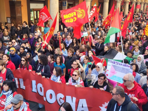 View of hundreds of people, a section of a giant march, filling an Italian street. Some marchers carry a huge red banner "FIOM-CGIL Bologna," some carry union flags or Palestinian flags, and one carries a handmade sign that says "Palestina Libera" with the flag and hearts. Mouths are open to sing or chant. Picturesque columns along the street. Marchers are diverse in race and gender.