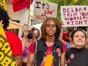 Several people with colorful signs in red and orange face the camera, one says “Delbar treat your workers right”
