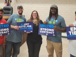 A group of five auto workers pose in their union hall with blue signs that say "End Tiers," "Every Auto Job a Good Job," and "United for a Strong Contract."