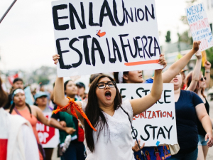 A group marches, one woman holds a sign above her head in Spanish: “En La Union Esta La Fuerza,” with a butterfly on it. Meaning “our strength lies in unity.”