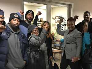 Nine Black film industry parking attendants in coats, smiling with fists up, indoors, in front of an NLRB office in New York City.