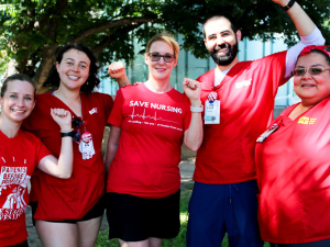 A group of five nurses in red shirts pose with their fists raised.
