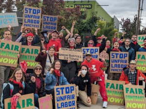 A group of workers and supporters hold cardboard signs reading Stop Unfair Labor Practices and Fair Contract Now. Many are wearing red bandanas.