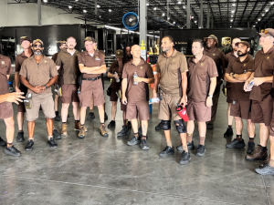 Fifteen workers in brown UPS uniforms stand in a semicircle while another uniformed worker speaks to them