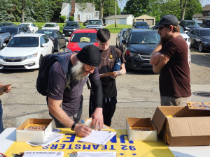A group of Teamsters in a parking lot stand around a table with petitions and leaflets on it. One is filling out a sign-up sheet.