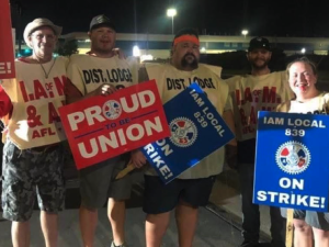 Four men and one woman wearing Machinists tabards and holding strike signs face the camera at night with plant lights behind them.