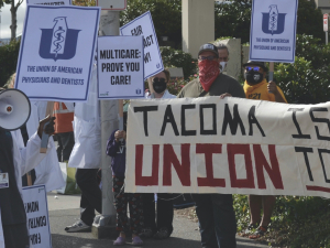 Urgent care doctors and staff in white coats picket alongside a sign held by union supporters that reads "Tacoma Is a Union Town."