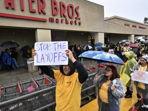Workers in yellow shirts and rain parkas picket in front of a grocery store with a ‘stop the layoffs’ signs visible