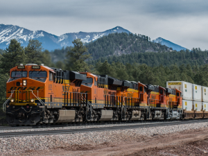 An orange BNSF train passes in front of a mountain and forested hills