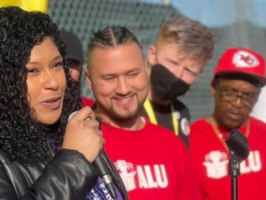 A woman speaks into a mic in foreground. Others wearing ALU T-shirts look on in background. The speaker and the listener nearest her are smiling. They are outdoors and it is sunny.