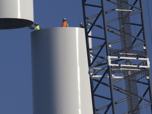 Workers join the top piece of a giant wind turbine to the bottom.  They are tiny against its hugeness.