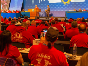 View from back of an auditorium at UAW convention. People in red shirts sit in rows; one red hoodie front and center has UAWD logo on the back. Onstage, Shawn Fain is at the podium, someone in a suit stands near him, and other people mostly in red shirts sit arrayed at long tables on the dais. A big blue screen behind them has UAW logo and intials repeated, and some other images not clearly visible.
