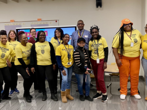 Fourteen workers, mostly Black and mostly women, pose smiling in a school classroom. Most are wearing yellow "Baltimore Teachers Union" T-shirts.