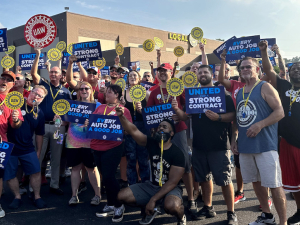 A fired up crowd of about 50 holds round yellow signs saying UAW Local 862 and blue and white signs saying United for a Strong Contract and Every Auto Job a Good Job.