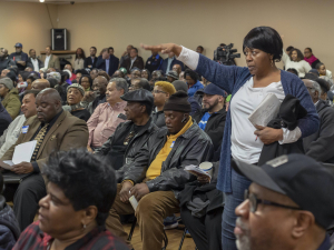 A woman stands up to speak in a big meeting, holding a notebook in one hand.