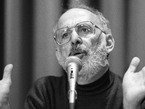 A bearded man speaks at a microphone with his hands wide open