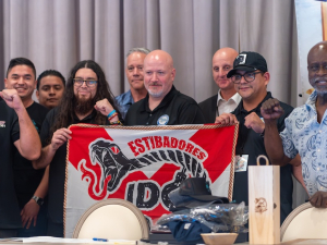 A group of a dozen men of various ages hold a red and white banner that says “Estibadores”