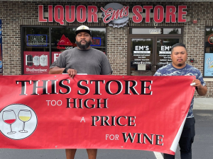 Three workers stand in front of a liquor store holding a banner that reads "This Store Too High a Price for Liquor"