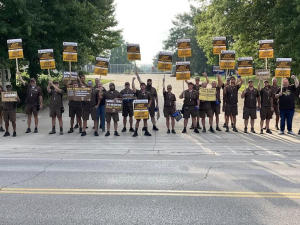 A row of people in UPS outfits with signs seen from far away block the road to some kind of facility, a gate is visible behind them.