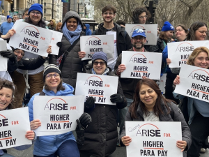About 15 smiling people hold identical signs saying “ARISE for Higher Para Pay”
