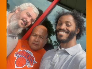 Three workers pose for a group selfie in front of the store. They read as three men of different races. The man in the middle is wearing an orange Knicks T-shirt.