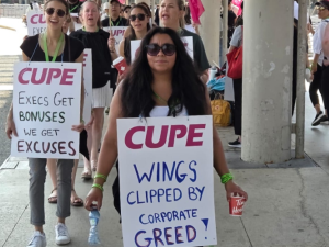 Flight attendants, mainly women, march on an airport sidewalk. Some wave pink CUPE flags and others wear sandwich-board signs with "CUPE" printed on and handwritten slogans. The two most visible say "Wings clipped by corporate greed!" and "Execs get bonuses, we get excuses." The woman in the foreground meets the camera's gaze with a serious expression; women behind her are chanting or singing.