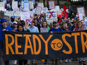 Big crowd marches behind a giant black banner hand-painted in orange "Ready to Strike." Many people hold handmade signs on sticks. A person near the front with long pink hair and reflecting sunglasses looks defiantly into the camera. A person in a blue beanie punches a tattooed arm into the air, raising a fist. Farther back, glimpse someone in a red inflatable lobster suit.