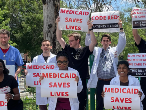 A diverse group of people, mostly in white lab coats, stand outside holding printed signs that say, in red on white, "Medicaid saves lives." Other signs say "Code red: patients in crisis" and "Our patients rely on Medicaid to get the care they need. This bill would end that."