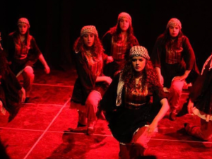 Seven women in long skirts and kerchiefs kneel, their arms in motion, apparently mid-dance. They are on a stage, bathed in red light.