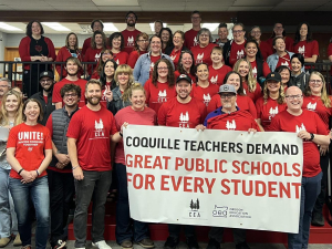 A group of dozens of teachers wearing red shirts gather behind a union banner.