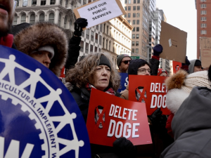 A crowd of people hold red and white signs saying “Delete Doge”