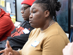 A Black woman in a tan shirt speaks while three men, white and Black, look on.