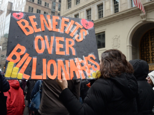 Marchers walk away from the camera with a giant orange on black sign, “Benefits over Billionaires”