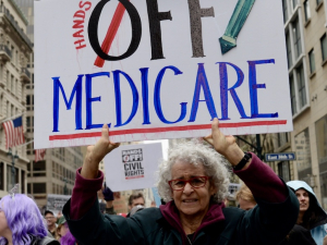 A woman in a big crowd holds up a sign saying ‘hands off Medicare’
