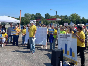 A group in a parking lot in yellow t-shirts listen while one man speaks through a microphone