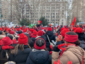 A sea of people, mostly women, of various races, bundled up in winter clothes with red hats and scarves, packs a square in New York City. One person with a megaphone stands elevated a bit in the center, surveying the crowd.