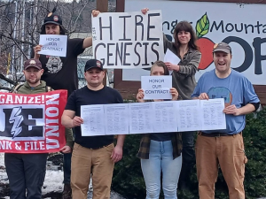 Eight people outside face the camera with signs, including “Hire Genesis” and a UE banner. 