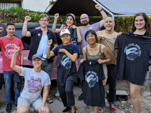 Eleven smiling people pose outside. Some are holding up silkscreened T-shirts with Tender Claws union logo showing a claw. Many are raising one fist, and one is pointing at another person in the group.