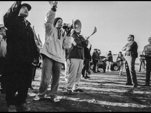 A vintage black-and-white photo shows a line of workers standing outside, one calling into a megaphone, facing down cops with folded arms. They are photographed from a low, dramatic angle.