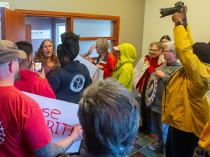 At least a dozen activists crowd up to the door of a VU Manufacturing office, where a company flack is gesuring widely and trying to fend them off. One activist is holding a camera aloft, others holding signs or wearing UAW or Labor Notes gear. A woman 