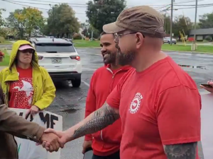 Screenshot shows Israel Cervantes on the left, in a weathered brown leather jacket, shaking hands with Sean Crawford, right, in a red UAW T-shirt and a baseball cap, while a few other activists carrying handmade signs look on approvingly. They're standing in a parking lot after a rain.